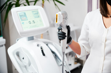 Close up of beautician hand in black glove holding ultrasound device for face lifting and skin tightening procedure. Young woman cosmetologist using modern cosmetology equipment in clinic.