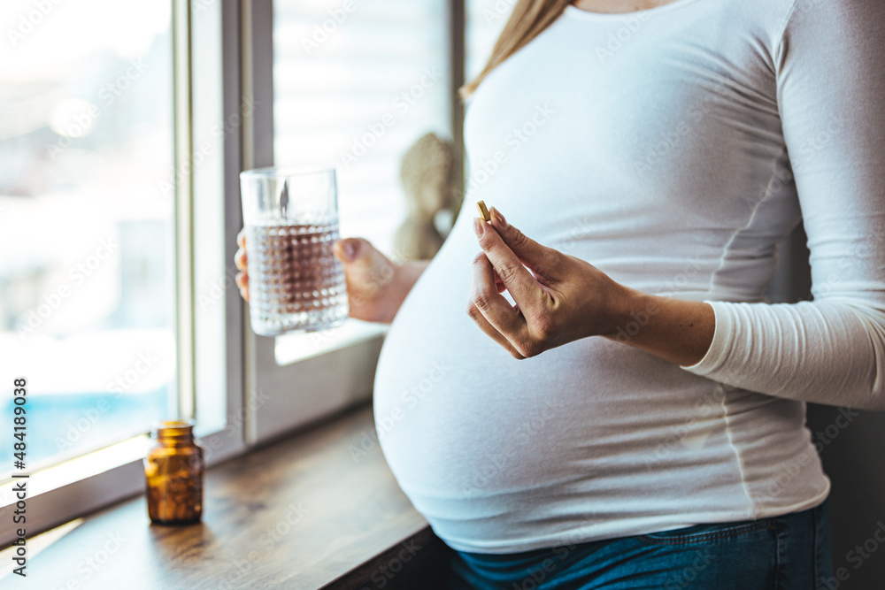 Pregnant woman reading label on bottle with medicine, with vitamins. Female sitting at home in bed with glass of water medicine. Pregnancy, health, pharmaceuticals, care and people.