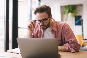 Wall Mural - Skilled hipster guy in optical eyewear browsing informative website on digital laptop technology using public internet connection in coworking space,Caucasian male programmer in glasses installing app