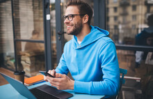 Portrait Of Cheerful Millennial Blogger With Modern Cellphone And Laptop Technology Enjoying Freelance Lifestyle, Happy Hipster Guy In Optical Eyewear Using Mobile Phone And Netbook In Street Cafe