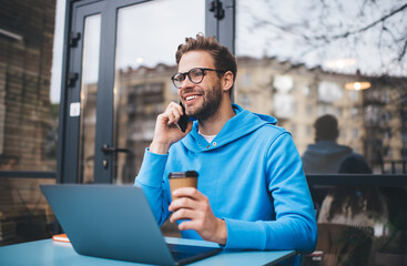 Wall Mural - Cheerful IT professional with laptop technology making smartphone conversation for discussing web programming, happy hipster guy in spectacles using modern netbook while calling via mobile app