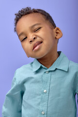 adorable child with dark skin, Cute playful young boy in shirt standing winking at camera, isolated on purple studio background. portrait of black child posing. childhood, human emotions concept