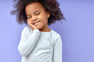 Happy girl kid keeps hands on cheeks,smiles satisfied,amiably,with eyes closed, has fun chatting with friends, wears casual clothes, isolated over purple background.Good mood concept.Copy space.