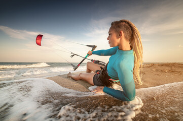 Wall Mural - Attractive caucasian woman kitesurfer in a neoprene wetsuit and with dreadlocks on her head is resting lying on a sandy beach on the shore holding her kite in the wind