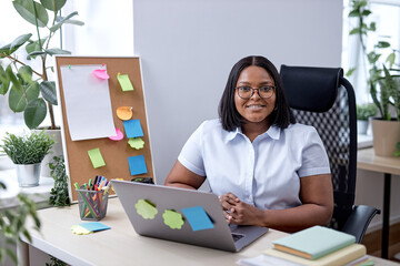 Wall Mural - Young business woman working on laptop at office, sitting behind office desk working on new project, smiling, feeling happy. beautiful black lady in formal wear. success and business concept