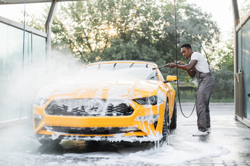 Poster - African stylish young focused man in work clothes washing his modern sport car with a water gun on self-service washing car station outdoors, rinsing the foam.