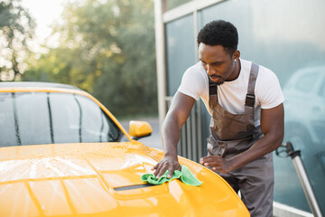 Wall Mural - Car wash and cleaning at outdoors self service station. Shot of handsome bearded young African guy worker cleaning yellow car hood with a green microfiber cloth outdoors in summer sunny morning