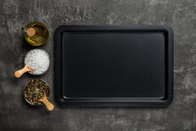 Empty baking sheet, olive oil jar, salt and pepper grains over black textured background. Rectangular oven tray and spices for baking and roasting. Baking pan for cooking and food design.