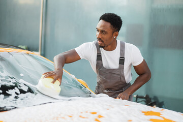 Canvas Print - Young attractive African American man washing his yellow luxury car in a self-service car wash station outdoors with cleaning foam and yellow sponge.