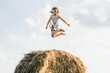 Portrait of little girl in dress playing and jumping on haystack in field. Light bright sunny day. Cheerful and enjoy freedom concept. High bounce. Reach sky. Side view. Low angle. One kid in field
