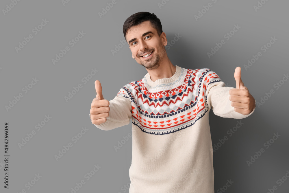 Handsome young man in knitted sweater showing thumb-up on grey background