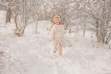 A Happy Little Girl In A White Knitted Hat And Sweater Runs Around A Snowy Park