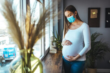 Happy Pregnant Woman wear medical face mask protect virus standing in front of windows stroking big belly with love at cozy home