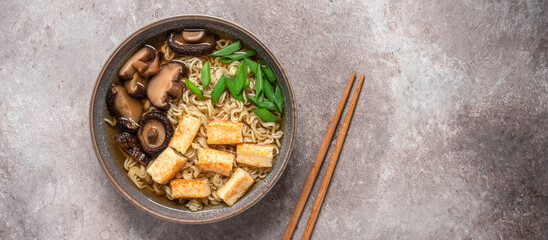 Asian vegan ramen noodle soup with roasted tofu cheese, chives and shiitake mushrooms in a bowl on brown grunge background. Top view. Selective Focus. Banner