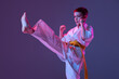 Portrait of sportive kid, male taekwondo, karate athletes in doboks doing basic movements isolated on purple background in neon. Concept of sport, martial arts