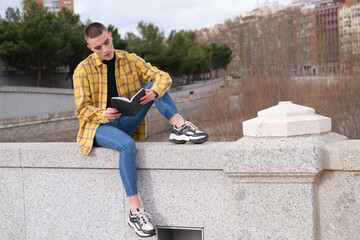 Wall Mural - Young non binary person sitting on a wall and reading a book.