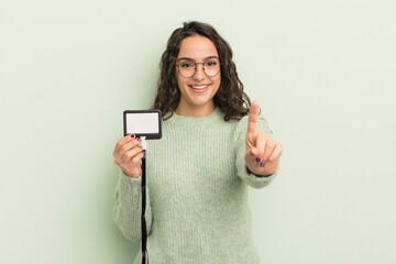 Wall Mural - young pretty hispanic woman smiling and looking friendly, showing number one. accreditation card id