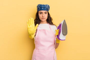 Wall Mural - young pretty hispanic woman looking serious showing open palm making stop gesture. iron and clothes concept