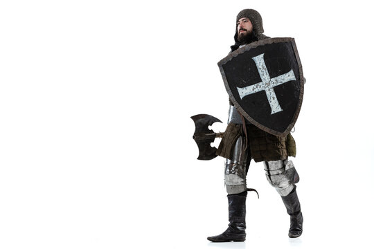 Full-length portrait of brave man, medieval knight, warrior with shield and ax calmly walking forward isolated over white studio background