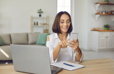 Woman using mobile phone. Happy beautiful lady in glasses sitting at desk with laptop, notebook and pen at home, holding cellphone, looking at screen, sending messages, or organizing business schedule