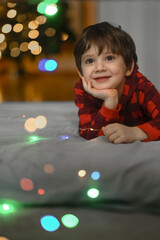 a five-year-old boy in red pajamas sits on the windowsill and looks out of the window among the New Year decorations