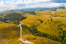 Wind Station With Powerful Machines Producing Clean Energy Operate On Hill Slopes Covered With Forests And Bushes Under Cloudy Sky Aerial View