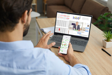 Very busy man working in home office, sitting at desk with laptop computer and mobile phone, organising business schedule, checking calendar, using synced devices, talking to colleague via videochat