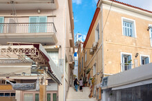 Two Women Climb The Stairs In A Narrow Alley Towards The Poros Clock Tower In The Old Town Center Of The Island Of Poros, Greece.