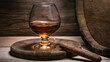 Cognac in glass next to cigar lies on wooden stand and old wooden barrel with cognac in wine cellar illuminated by soft light in rustic style.