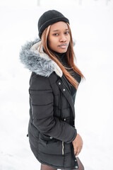 Wall Mural - beautiful young African model woman in fashionable clothes with a stylish winter jacket and hat walks outdoors with snow. Fashion pretty girl on a white background