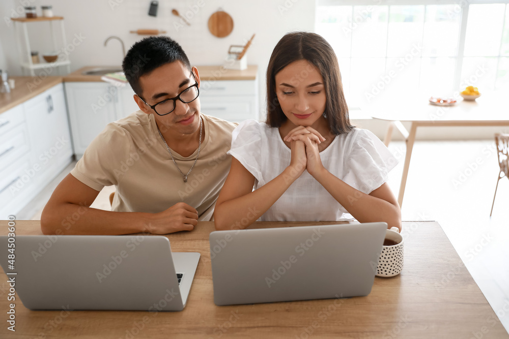 Young woman with her husband using laptops at table in kitchen