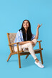 © Pixel-Shot - Beautiful young woman in wooden armchair pointing at something on blue background