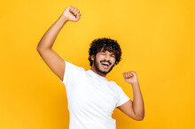 Ecstatic Lucky Happy Curly Indian Man, Dancing, Gesturing With Fists, Receives Profit, Glad To Win Lottery, Standing On Isolated Orange Background. Good News, Success And Happiness Concept