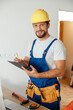 © Kostiantyn - Joyful handyman worker, builder in uniform and hard hat looking at camera, making notes on a clipboard while standing indoors, getting ready for renovation work