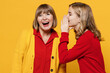 Happy woman 50s in red shirt have fun with teenager girl 12-13 years old. Grandmother granddaughter whispering gossip and tells secret behind her hand sharing news isolated on plain yellow background