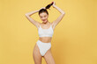 © ViDi Studio - Smiling lovely pretty nice attractive young brunette woman 20s wearing white underwear with perfect fit body standing ties hair in ponytail isolated on plain yellow color background studio portrait