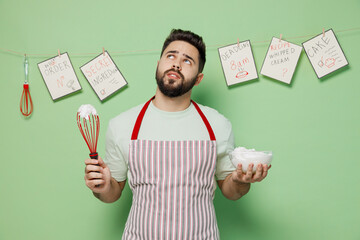 Wall Mural - Young puzzled minded male chef confectioner baker man 20s in striped apron whisk eggs for cream cake or omelette isolated on plain pastel light green background studio portrait Cooking food concept.