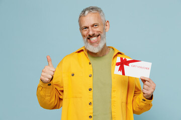 Elderly gray-haired mustache bearded man 50s wear yellow shirt hold store gift certificate coupon voucher card show thumb up isolated on plain pastel light blue background. People lifestyle concept.