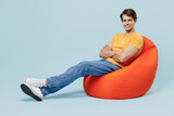Fototapeta Panele - Full body smiling happy young man 20s in yellow t-shirt sit in bag chair hold hands crossed folded look camera isolated on plain pastel light blue background studio portrait. People lifestyle concept.