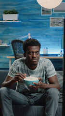 Wall Mural - Black adult watching comedy movie on television while eating popcorn from bowl. African american person laughing and looking at TV with snack. Man of african ethnicity having fun