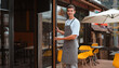 © shangarey - barista or waiter cafe or coffee shop owner against entrance, gesture inviting you to visit, smiling guy in apron standing outdoors being proud of his small local business