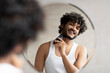 © Prostock-studio - Grooming concept. Handsome young indian man looking at mirror and shaving beard with trimmer or electric shaver