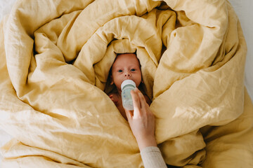 Wall Mural - Mother bottle feeding baby lying in bed covered with big yellow blanket, top view.