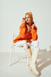 Fashion portrait of confident blonde woman wearing trendy orange sweatshirt, white skinny jeans, high leather boots, posing on white background. 