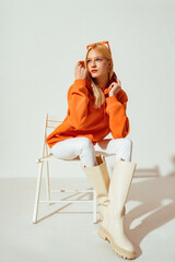 Wall Mural - Fashion portrait of confident blonde woman wearing trendy orange sweatshirt, white skinny jeans, high leather boots, posing on white background. 