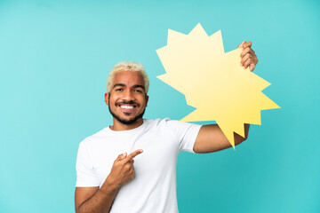 Young Colombian handsome man isolated on blue background holding an empty speech bubble and pointing it