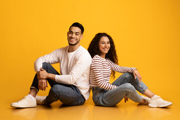 Wall Mural - Cheerful Young Arab Couple Sitting Back To Back Over Yellow Studio Background