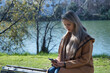 © Manuel - Young blonde woman, sitting on top of a bench, in a park, consulting social networks on her cell phone. Concept smartphone, social networks, connectivity, addiction.
