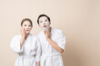 © ME Image - Happy Asian couple in bathrobe or spa suit doing sheet mask on face for facial treatment together on brown isolated studio background.