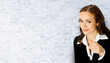 © vgstudio - Smiling business woman in black suit pointing forefinger at viewer, copy space for text. Brunette businesswoman, on white bricks loft wall background